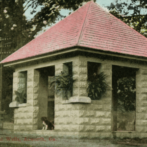 A small stone pavilion with a red hipped roof, arched openings, and hanging plants. A dog sits at the entrance. Text at bottom reads “Wells, Acworth, Ga.” Learn More About Historic Acworth Locations.
