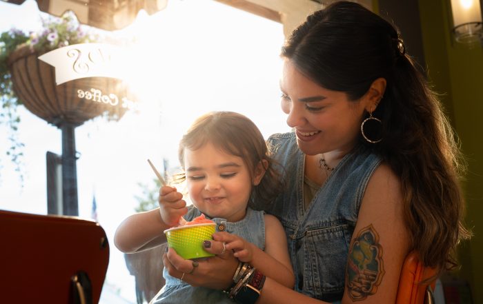 A woman smiles while helping a young child eat from a green bowl at a cafe, sunlight streaming through the window behind them—just one of the many ways to stay cool this summer in Acworth.