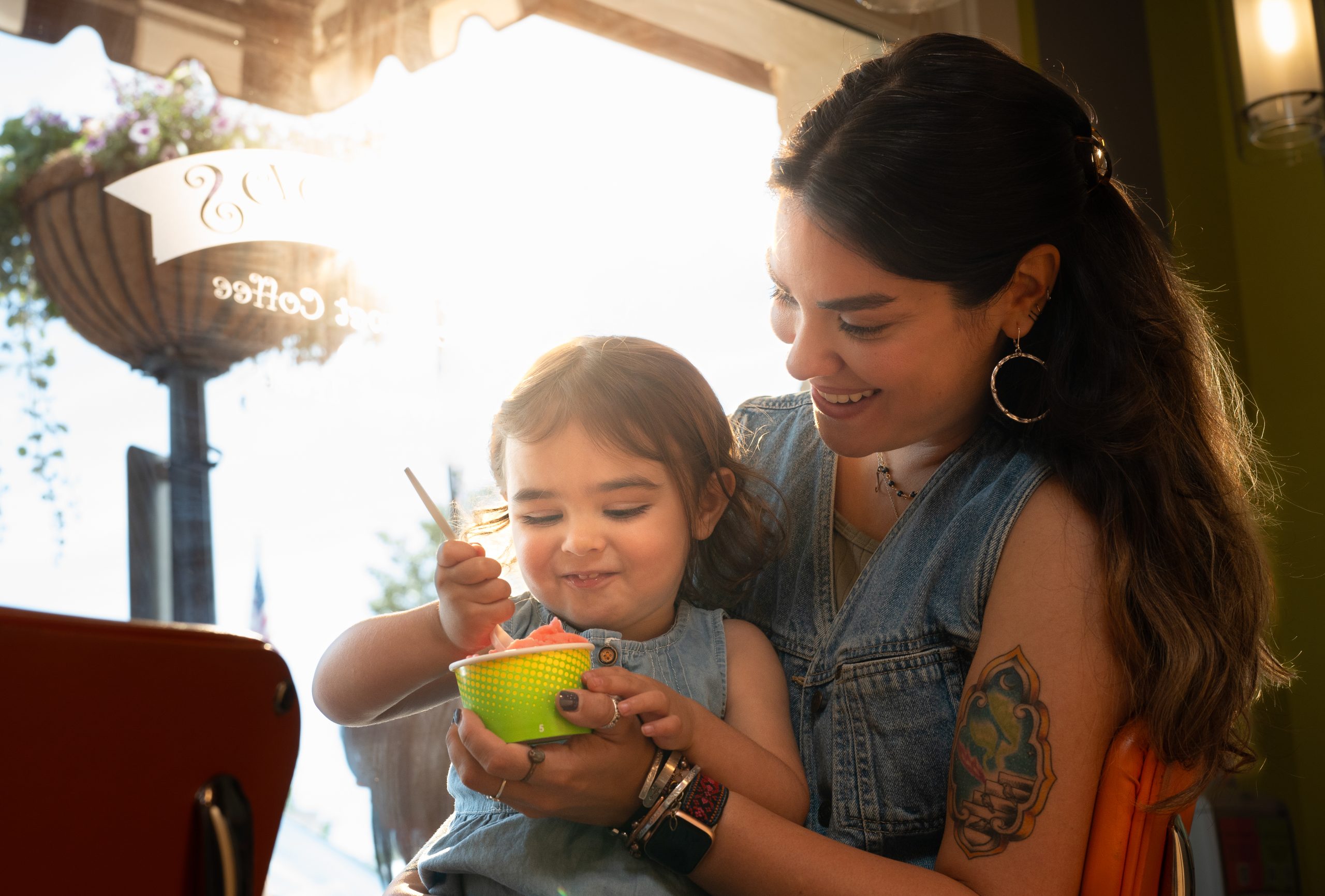 A woman smiles while helping a young child eat from a green bowl at a cafe, sunlight streaming through the window behind them—just one of the many ways to stay cool this summer in Acworth.