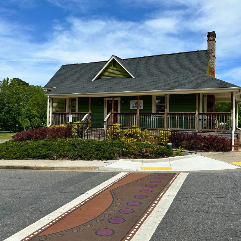 A green house with a wraparound porch sits by a street with a crosswalk painted in brown and purple circles; trees and bushes surround the property. Learn More About Historic Acworth Locations nearby. A green house with a wraparound porch sits by a street with a crosswalk painted in brown and purple circles; trees and bushes surround the property. Learn More About Historic Acworth Locations nearby.