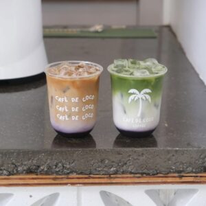 Two iced drinks in plastic cups with "Café de Coco" branding sit on a dark countertop; one is brown and purple, the other green, white, and purple—perfect to help you stay cool in Acworth.