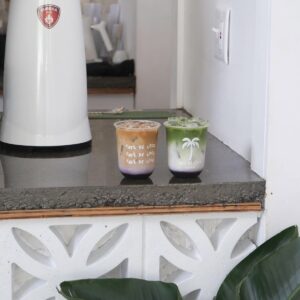 Two takeaway drinks, one iced coffee and one matcha, sit on a concrete counter near a white coffee machine in a minimalistic café—perfect for anyone looking to stay cool in Acworth.