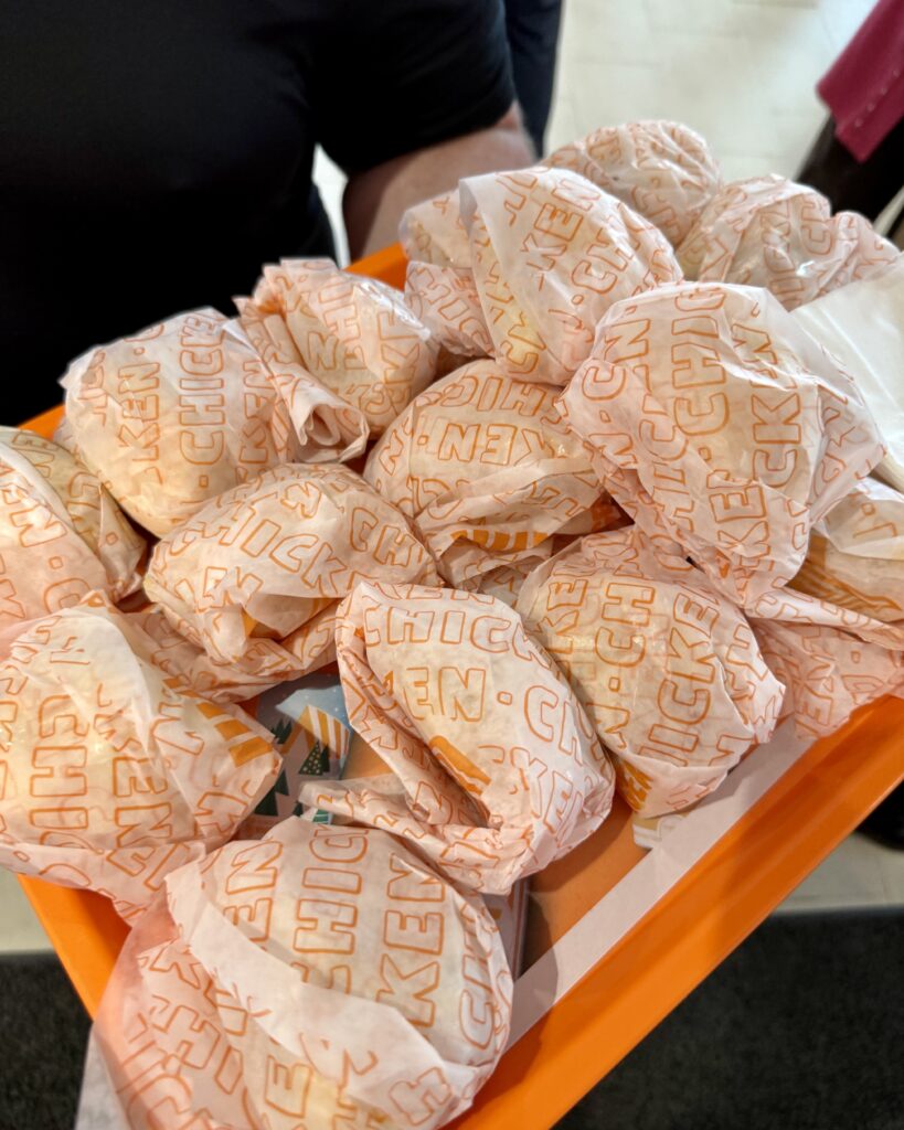 A person holds a tray filled with several wrapped chicken sandwiches in orange and white paper labeled 