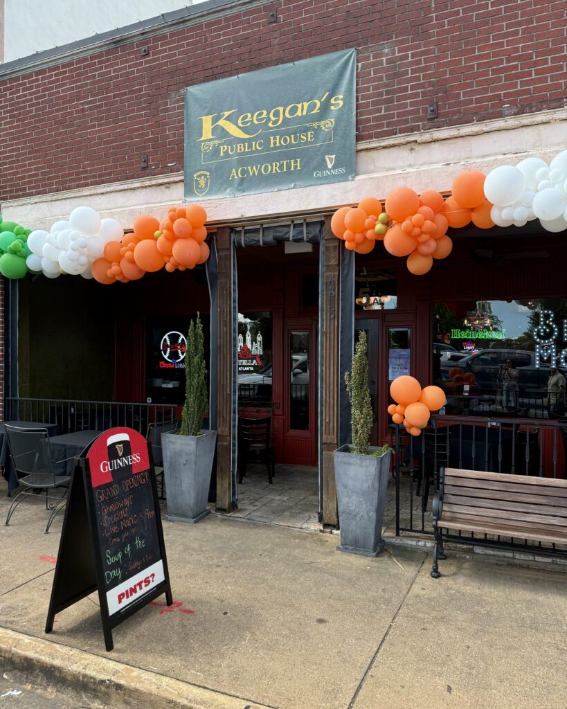 Keegan’s Public House exterior decorated with orange, white, and green balloons; sidewalk sign advertises a grand opening and pint special; benches and plants outside.