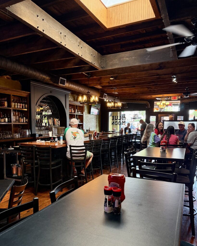 A bar and restaurant interior with wooden beams, bar stools, and tables. A few patrons are seated at the bar and a group stands near the entrance. Natural light enters through a skylight.