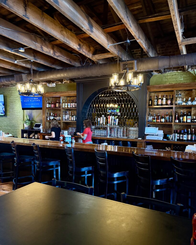 A bar with wooden beams on the ceiling, shelves of liquor bottles, several beer taps, barstools, and two people working behind the counter.