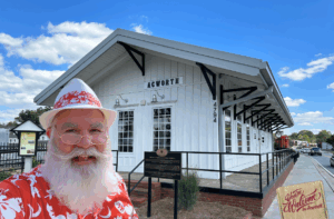 A man with a white beard and hat poses in front of the historic Acworth train depot building on a sunny day. A "Welcome to Acworth" sign is in the foreground.