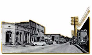 Black and white photo of a small town street with vintage cars, brick buildings, utility poles, and storefronts lining both sides of the road.