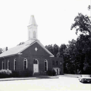 Black-and-white photo of a brick church with a steeple, arched windows, and an entrance ramp; a car is parked nearby beside trees. Learn More About Historic Acworth Locations captured in timeless detail.