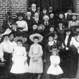 A black and white photo shows a large group of men, women, and children in formal attire posing in front of a brick building. Learn more about historic Acworth locations connected to moments like this.