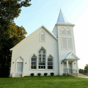 White church building with tall steeple, arched windows, and a ramp entrance, surrounded by green grass and trees under a clear sky. Learn more about historic Acworth locations like this charming landmark.
