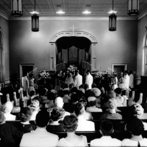 Black and white photo of a wedding ceremony in a church, with the bride and groom at the altar, surrounded by their wedding party and guests. Learn more about historic Acworth locations for timeless celebrations like this one.