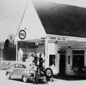 A vintage black-and-white photo of a gas station labeled “Hughes Ser. Sta.” with a “PURE” sign. A person is refueling a classic car at the pumps. Learn more about historic Acworth locations through this nostalgic scene.