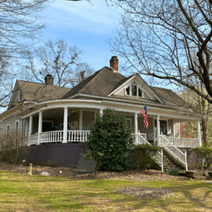 A gray, one-story house with a wraparound porch and an American flag on the railing, surrounded by trees in early spring. Learn more about historic Acworth locations with charming homes like this.