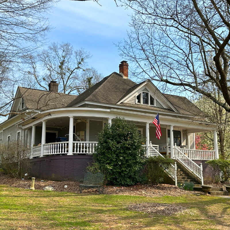 A gray, one-story house with a wraparound porch and an American flag on the railing, surrounded by trees in early spring. Learn more about historic Acworth locations with charming homes like this. A gray, one-story house with a wraparound porch and an American flag on the railing, surrounded by trees in early spring. Learn more about historic Acworth locations with charming homes like this.