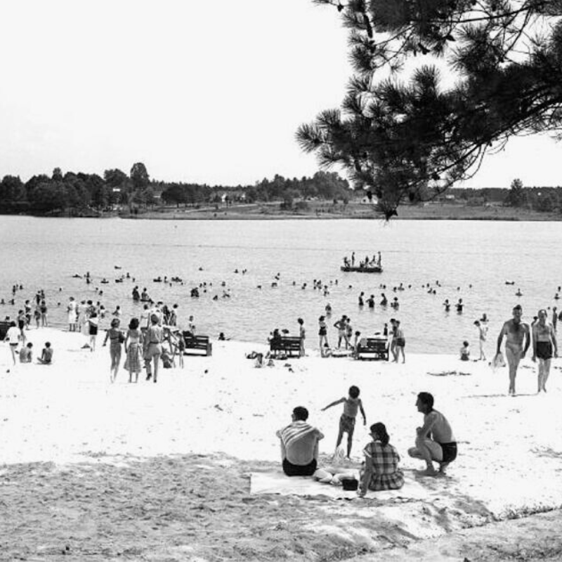 Black and white photo of people relaxing on a sandy beach and swimming in a lake, with trees and benches in the background. Learn more about historic Acworth locations captured in moments like these. Black and white photo of people relaxing on a sandy beach and swimming in a lake, with trees and benches in the background. Learn more about historic Acworth locations captured in moments like these.