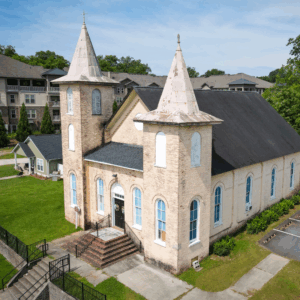 Aerial view of a historic brick church with two steeples, arched windows, and a black roof, surrounded by grass and nearby buildings. Learn more about historic Acworth locations like this architectural treasure.
