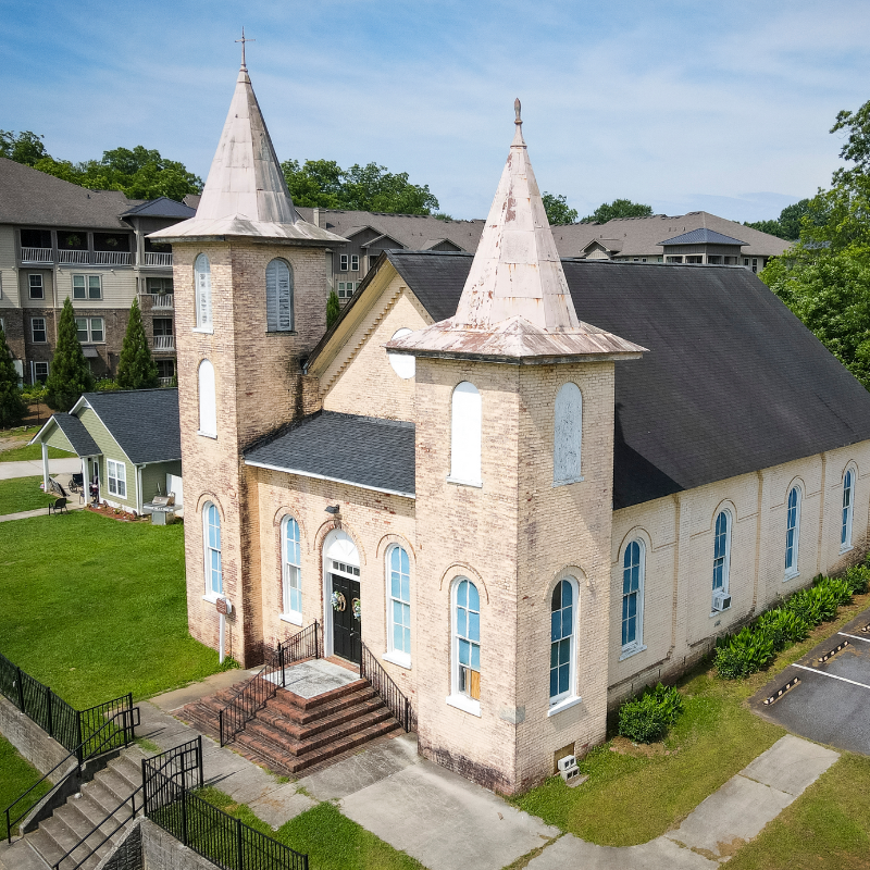 Aerial view of a historic brick church with two steeples, arched windows, and a black roof, surrounded by grass and nearby buildings. Learn more about historic Acworth locations like this architectural treasure. Aerial view of a historic brick church with two steeples, arched windows, and a black roof, surrounded by grass and nearby buildings. Learn more about historic Acworth locations like this architectural treasure.