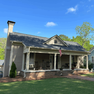 A single-story house with a covered front porch, rocking chairs, an American flag, brick steps, and a well-kept lawn under a clear blue sky. Learn more about historic Acworth locations like this charming home.