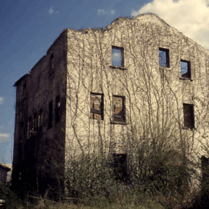 An abandoned, three-story building with boarded windows and vines climbing its weathered exterior under a partly cloudy sky. Learn More About Historic Acworth Locations as you discover stories hidden within these timeless walls.