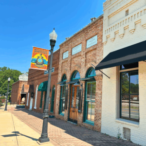 Brick buildings with arched windows and awnings line a sunny street in a historic downtown area, with a streetlamp and a "Historic Downtown" banner in the foreground. Learn more about Historic Acworth locations as you explore this charming scene.