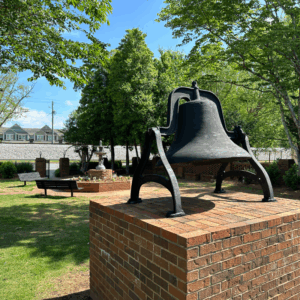 A large metal bell is mounted on a brick pedestal in a shaded outdoor park with trees, benches, and a fountain in the background. Learn More About Historic Acworth Locations while enjoying this peaceful setting.