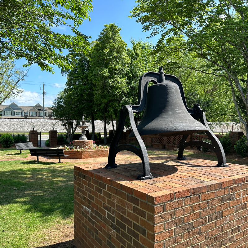 A large metal bell is mounted on a brick pedestal in a shaded outdoor park with trees, benches, and a fountain in the background. Learn More About Historic Acworth Locations while enjoying this peaceful setting. A large metal bell is mounted on a brick pedestal in a shaded outdoor park with trees, benches, and a fountain in the background. Learn More About Historic Acworth Locations while enjoying this peaceful setting.