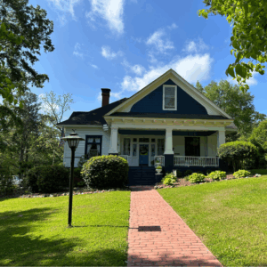 A brick path leads to a blue and white house with a front porch, surrounded by green lawn, bushes, and trees under a clear sky. Learn more about historic Acworth locations like this charming residence.