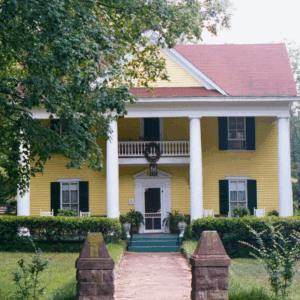 A two-story yellow house with white columns, green shutters, and a front porch, surrounded by trees and greenery—learn more about historic Acworth locations like this charming home.