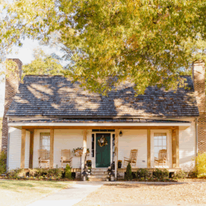 A small white house with a shingled roof, green front door, and two rocking chairs on the porch sits among autumn trees—inviting you to learn more about historic Acworth locations.