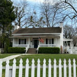 A white, single-story house with a front porch, American flag, picket fence, and green yard, surrounded by trees and under a partly cloudy sky. Learn more about historic Acworth locations in this charming setting.
