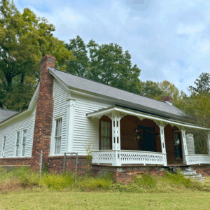 A white wooden house with a brick foundation, decorative front porch, and large brick chimney stands among trees and grass under a cloudy sky. Learn more about historic Acworth locations like this charming home.
