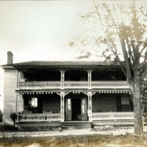 Two-story wooden house with ornate railing on both levels, a covered porch, and a large tree in the front yard. The house appears old and weathered—learn more about historic Acworth locations like this charming home.