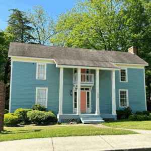 A two-story blue house with white trim, a small second-floor balcony, four white columns, and a peach front door stands among green bushes. Learn more about Historic Acworth Locations like this charming home.