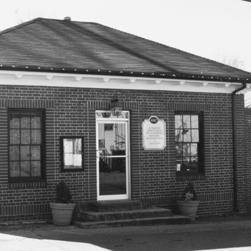 A small, single-story brick building with two windows, a central door, two potted plants, and a plaque beside the entrance. Learn more about historic Acworth locations when you visit this charming site. A small, single-story brick building with two windows, a central door, two potted plants, and a plaque beside the entrance. Learn more about historic Acworth locations when you visit this charming site.