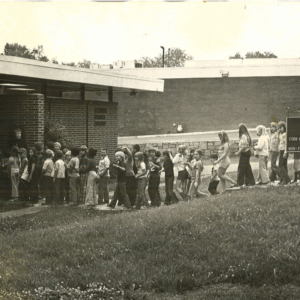 A group of children and a few adults stand in line outside a brick building, approaching the entrance; a sign is partially visible on the right. Learn more about historic Acworth locations as you explore this site.