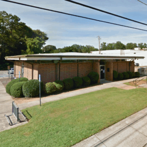 A single-story brick building with trimmed bushes lining the front, a sidewalk, bike rack, and a grassy lawn under a partly cloudy sky. Learn more about historic Acworth locations as you explore this charming spot.