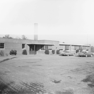 Black and white photo of a single-story brick building with a chimney, surrounded by parked vintage cars and sparse landscaping. Learn More About Historic Acworth Locations captured in this timeless scene.