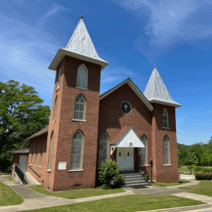 Red brick church with two white steeples, arched windows, and front steps, set against a blue sky with trees—Learn More About Historic Acworth Locations.