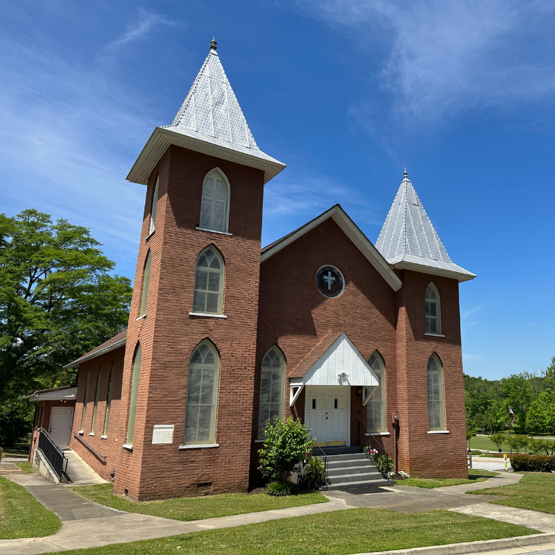 Red brick church with two white steeples, arched windows, and front steps, set against a blue sky with trees—Learn More About Historic Acworth Locations. Red brick church with two white steeples, arched windows, and front steps, set against a blue sky with trees—Learn More About Historic Acworth Locations.