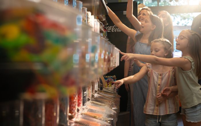 A woman and three children stand in front of a candy display, each pointing at different bins filled with various colorful candies—checking off a sweet treat from their end of summer bucket list.