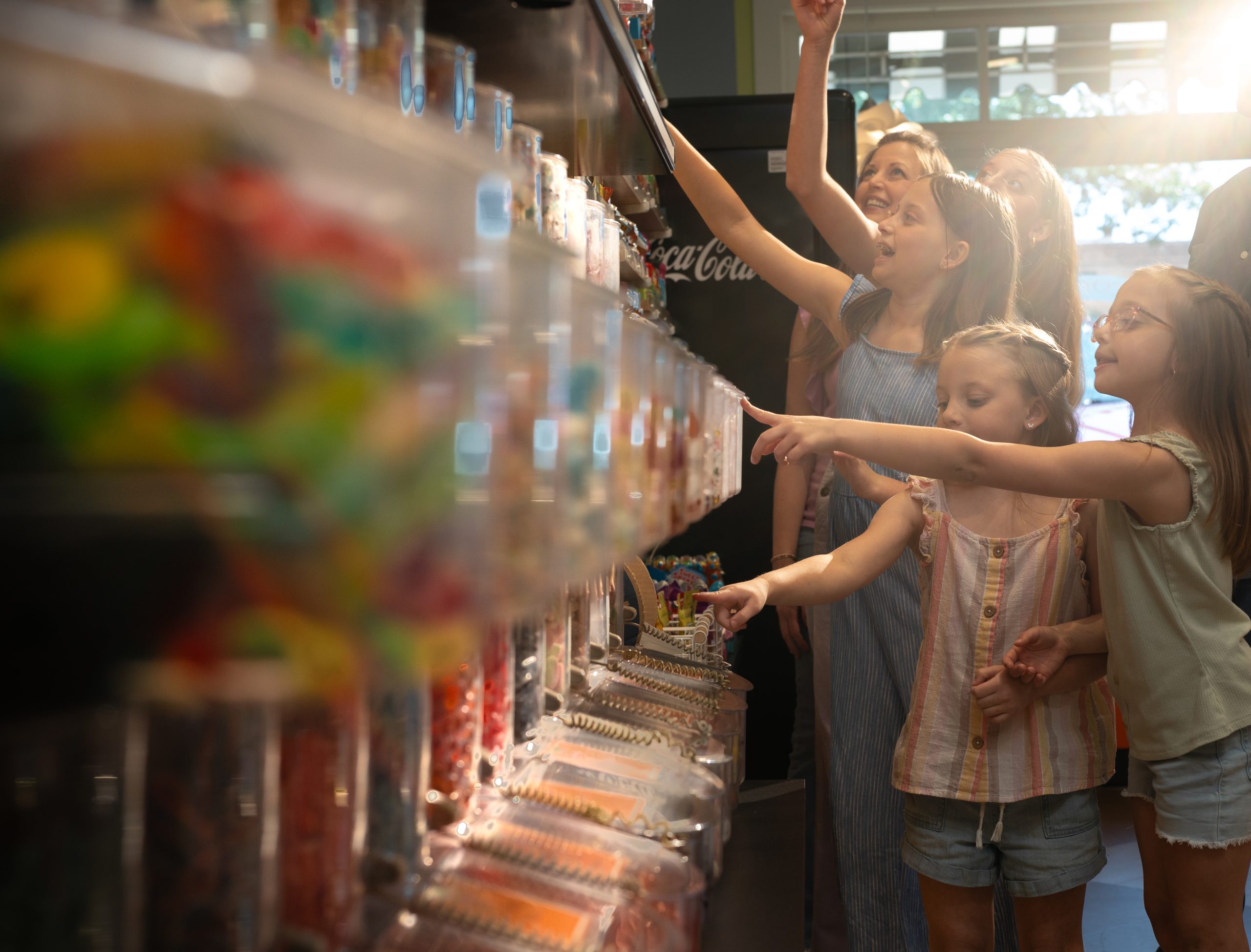 A woman and three children stand in front of a candy display, each pointing at different bins filled with various colorful candies—checking off a sweet treat from their end of summer bucket list.