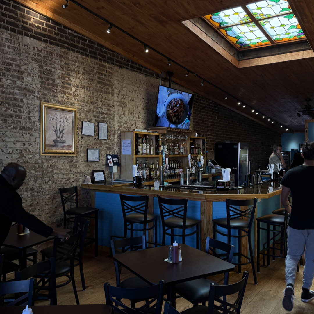 A bar area in a restaurant with wooden tables and chairs, exposed brick walls, a stained glass ceiling panel, and a TV above the bar—perfect for your end of summer bucket list gathering. A bar area in a restaurant with wooden tables and chairs, exposed brick walls, a stained glass ceiling panel, and a TV above the bar—perfect for your end of summer bucket list gathering.