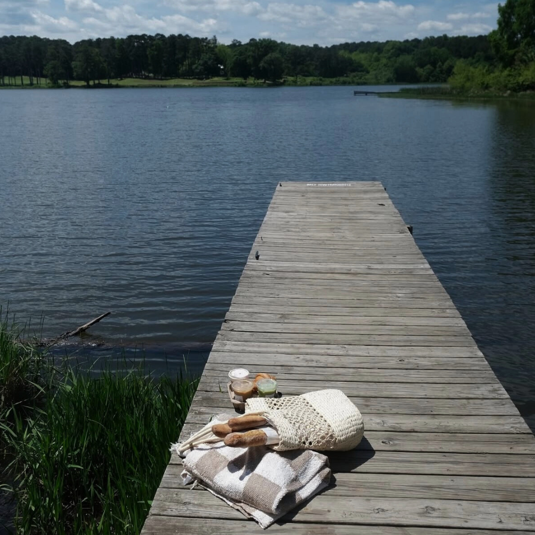A wooden dock extends over a calm lake with a picnic setup—bread, drinks, and a blanket—placed near the edge. Trees and grass surround the water, making it perfect for your end of summer bucket list. A wooden dock extends over a calm lake with a picnic setup—bread, drinks, and a blanket—placed near the edge. Trees and grass surround the water, making it perfect for your end of summer bucket list.