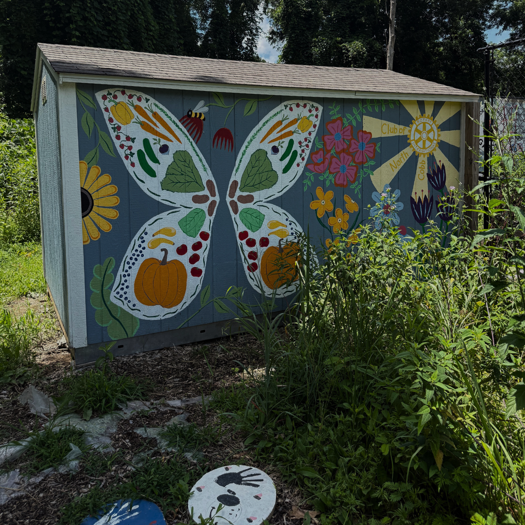 A shed with a mural featuring a large butterfly, flowers, and a sun stands among overgrown grass and plants, capturing an end of summer bucket list vibe in a lush garden setting. A shed with a mural featuring a large butterfly, flowers, and a sun stands among overgrown grass and plants, capturing an end of summer bucket list vibe in a lush garden setting.