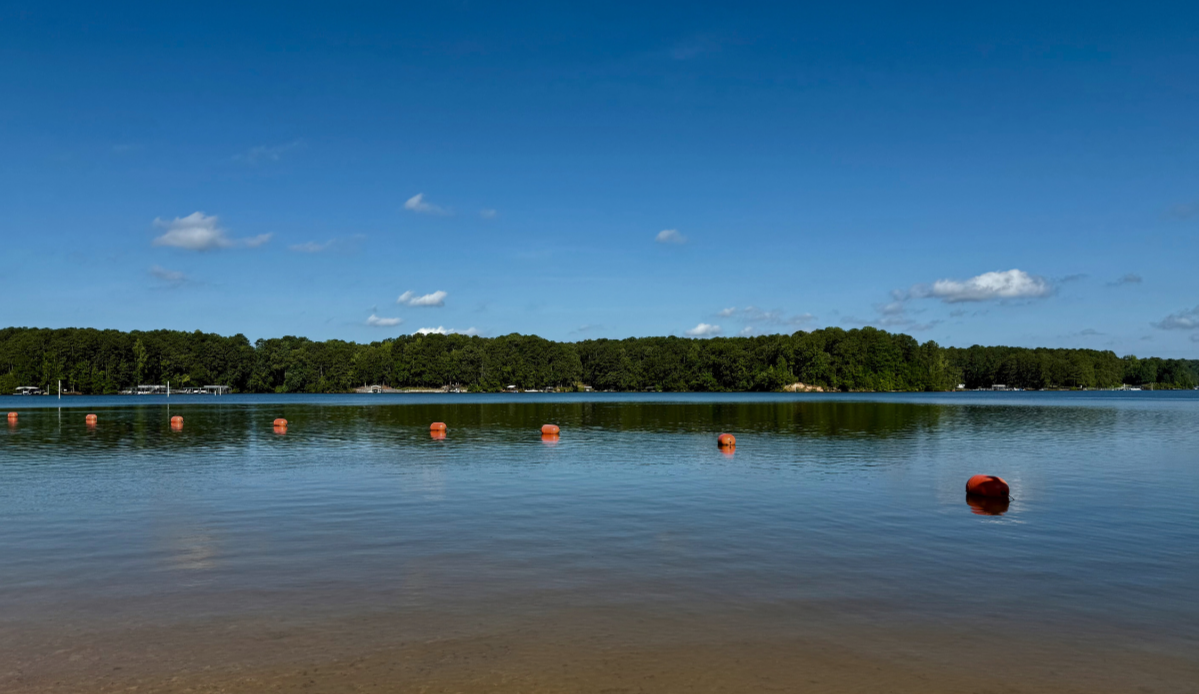 A calm lake with orange buoys floating in a line near the shore, bordered by a forested area under a blue sky with a few clouds—a perfect spot to check off your end of summer bucket list. A calm lake with orange buoys floating in a line near the shore, bordered by a forested area under a blue sky with a few clouds—a perfect spot to check off your end of summer bucket list.