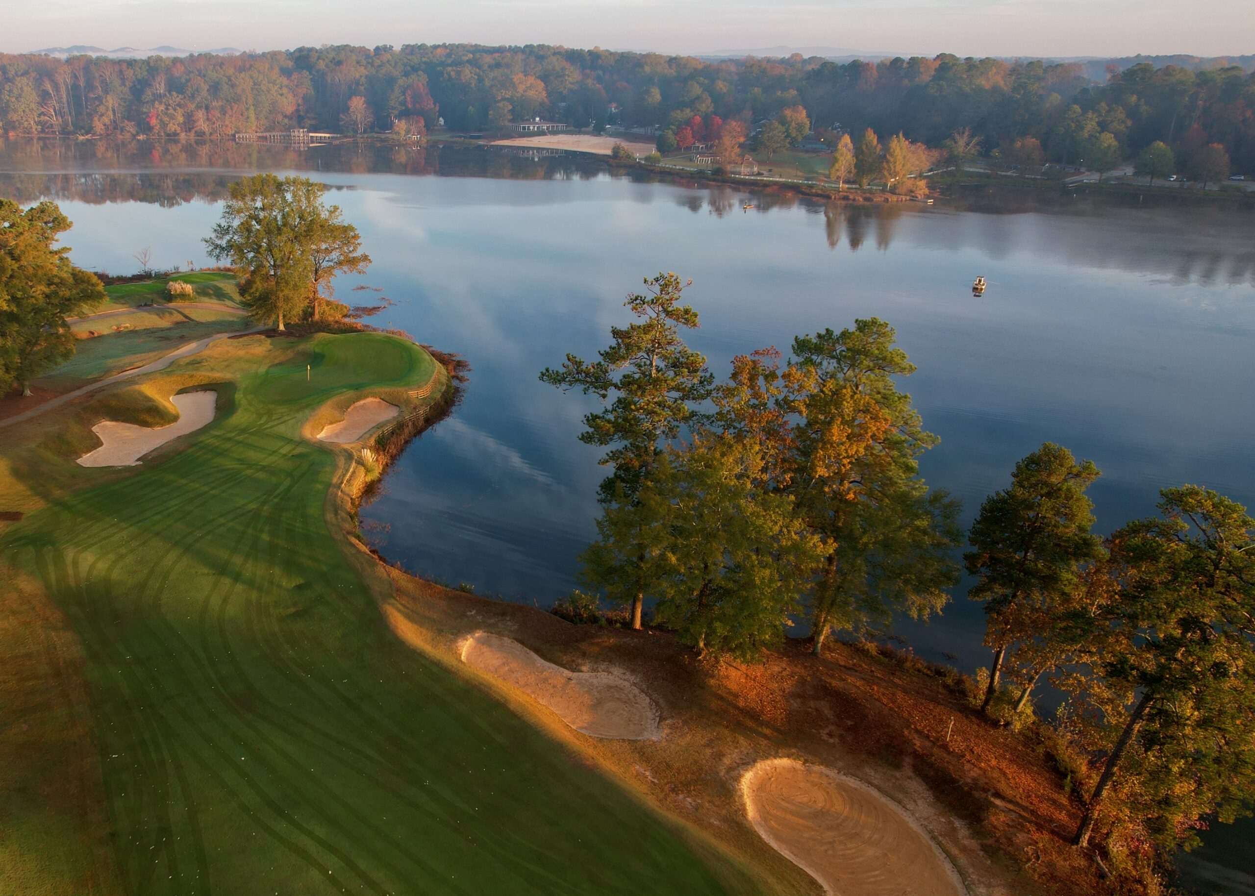 Aerial view of a golf course next to a calm lake, with sand bunkers, trees, and fall foliage reflected in the water.