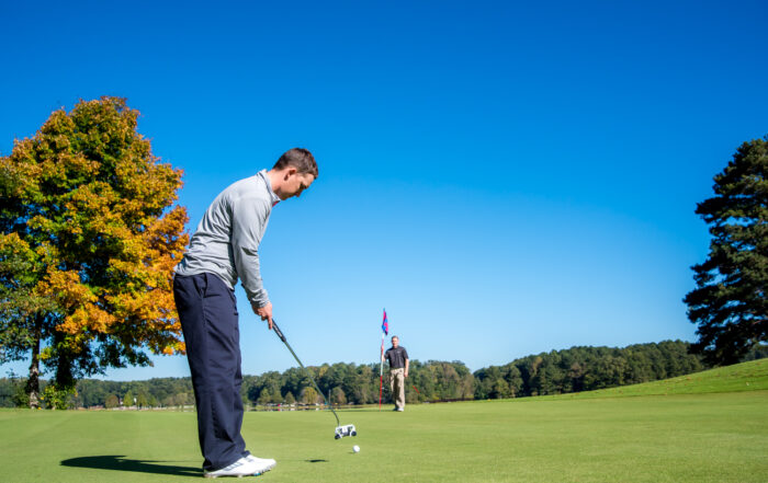 A man in a gray shirt and dark pants putts a golf ball on a green, while another person stands near the flag in the background under a clear blue sky.