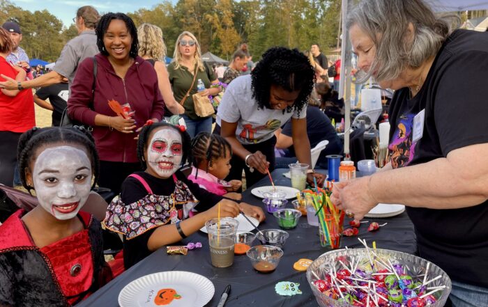 Children with painted faces sit at a table with crafts and candy, while adults assist and others stand nearby during HALLOWEEN IN ACWORTH's festive outdoor event.