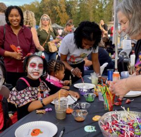 Children with painted faces sit at a table with crafts and candy, while adults assist and others stand nearby during HALLOWEEN IN ACWORTH's festive outdoor event.